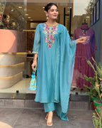 Woman in a blue traditional outfit standing in front of a store window with another outfit displayed.